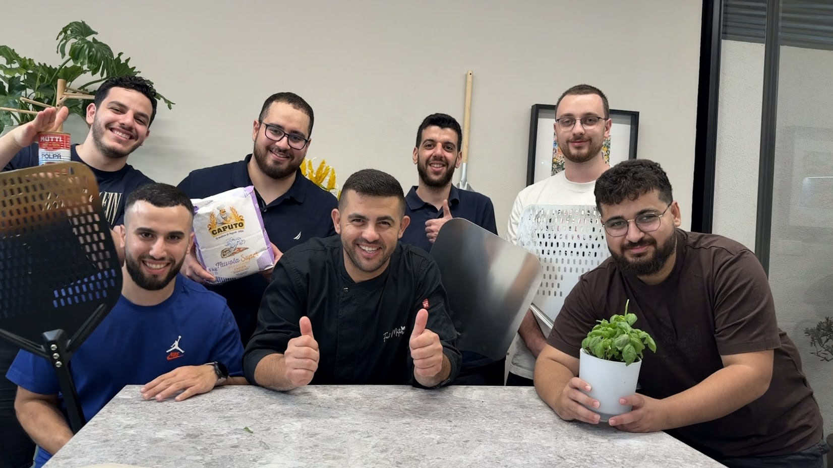 مجموعة من المشاركين بورشة البيتزا مع الشيف حول طاولة المطبخ، معهم أطراف البيتزا وطحين